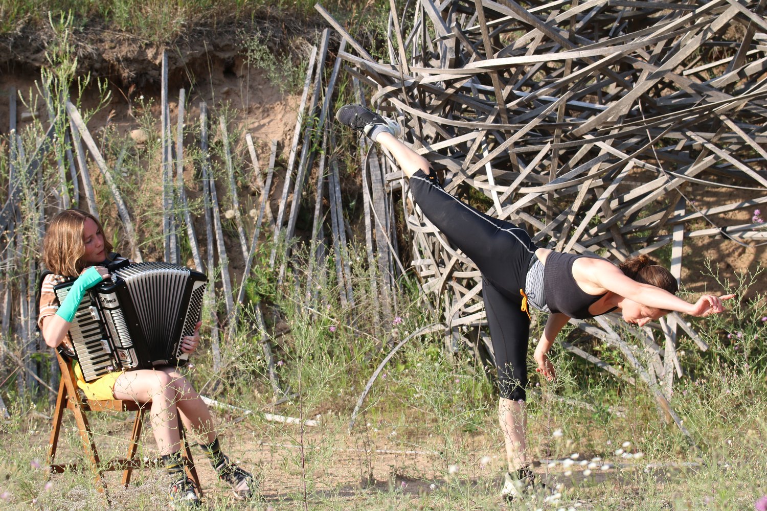 Kiera plays accordion and sings while Ella extends one leg and two arms in opposition in front of a large, wooden, nest-like sculpture. They are surrounded by scratchy star thistle plants and dry dirt.